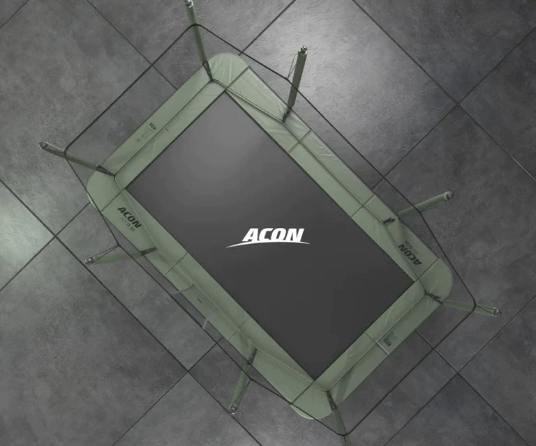 Bild på rektangulär Acon HD-studsmatta tagen från fågelperspektiv, mot grå bakgrund. Studsmattans svarta matta har vit ACON-logotyp och gröna kantskydd 