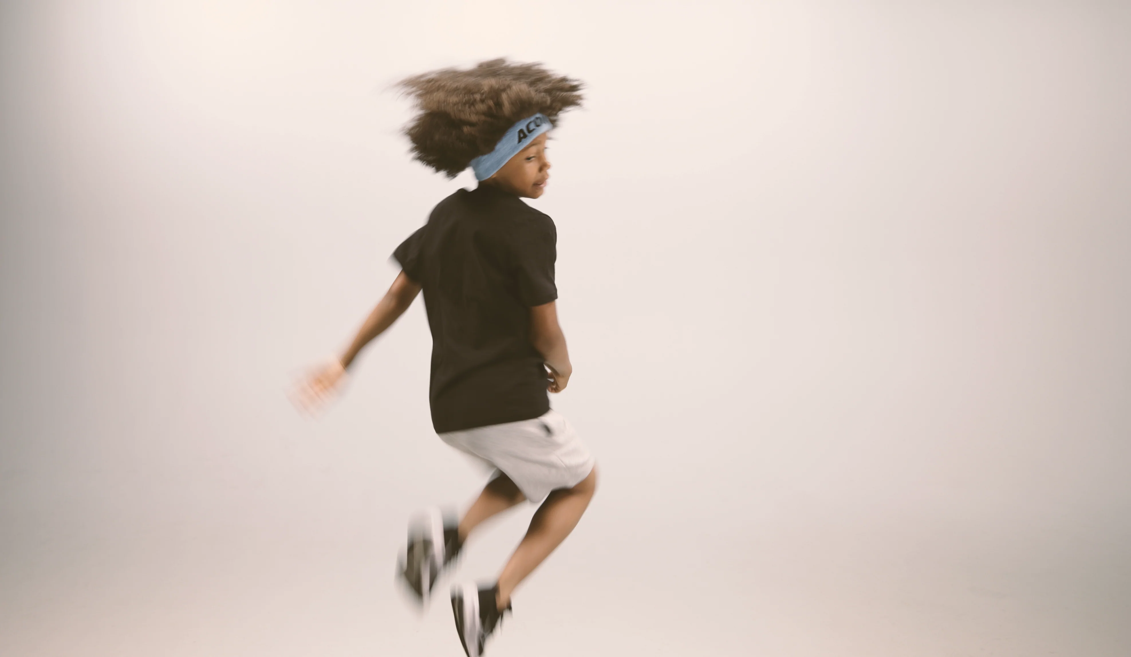 Ett hoppande barn med Acon-pannband, svart T-shirt, vita shorts och sneakers. Bilden är tagen mot beige bakgrund 