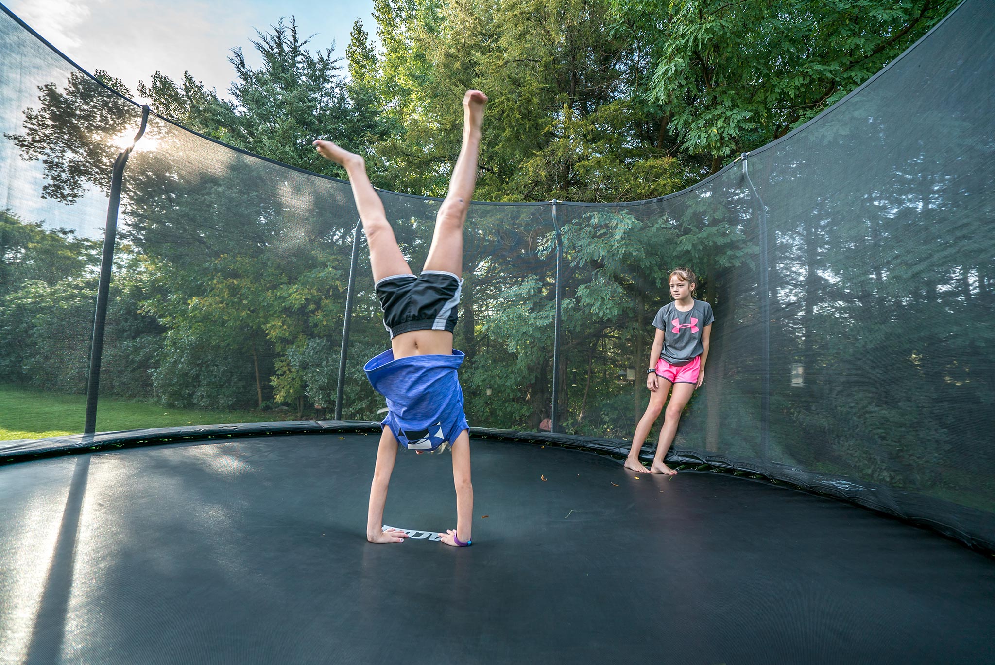 En flicka som står på trampolin med händerna, en kamrat som lutar sig mot säkerhetsnätet och tittar på.